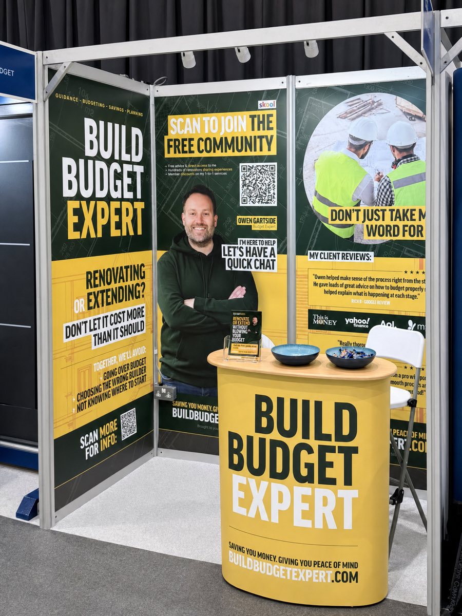 The full Build Budget Expert exhibition stand assembled at a trade show: matching dark green and gold roller banners, pull-up displays and a branded podium
