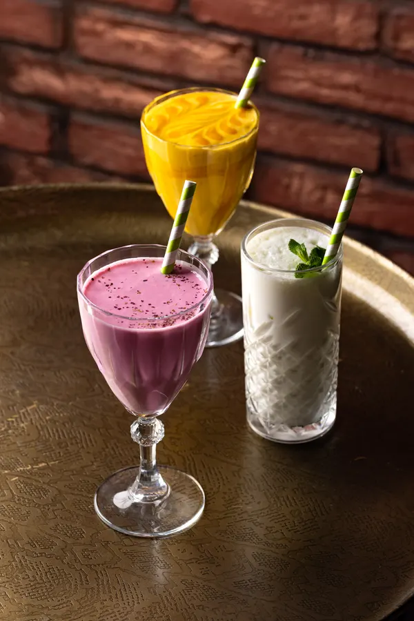 A mango lassi and a berry lassi beside a white drink with mint, against a brick wall
